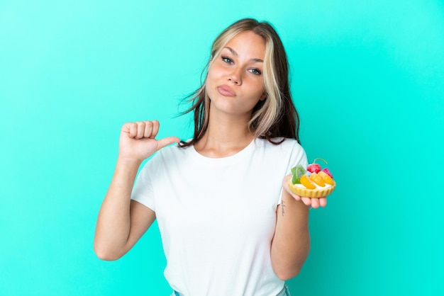 Jovem russa segurando um doce de fruta isolado em fundo azul orgulhoso e satisfeito