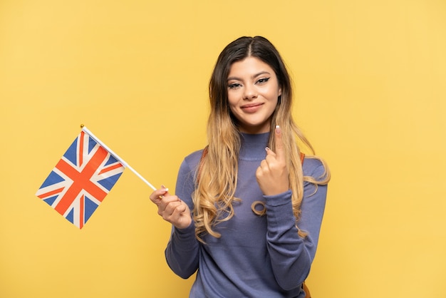 Jovem russa segurando uma bandeira do Reino Unido, isolada em um fundo amarelo, fazendo um gesto de aproximação