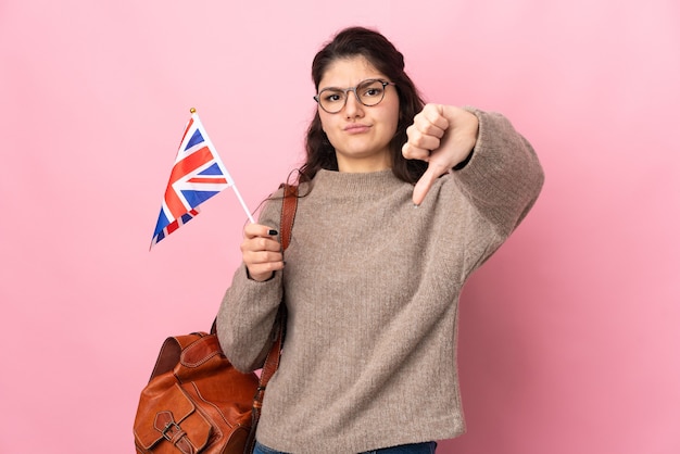 Jovem russa segurando uma bandeira do reino unido isolada em um fundo rosa, mostrando o polegar para baixo com expressão negativa