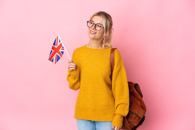 Jovem russa segurando uma bandeira do Reino Unido isolada na parede rosa, olhando para o lado e sorrindo