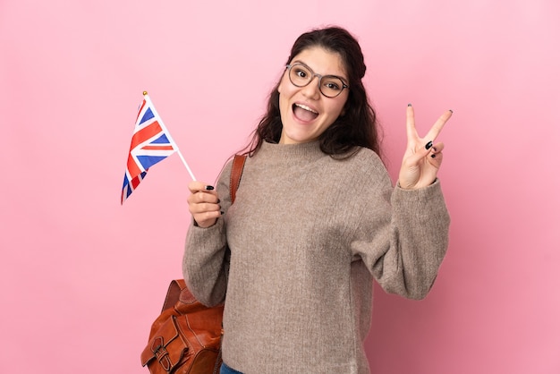 Jovem russa segurando uma bandeira do Reino Unido isolada na parede rosa, sorrindo e mostrando o sinal da vitória