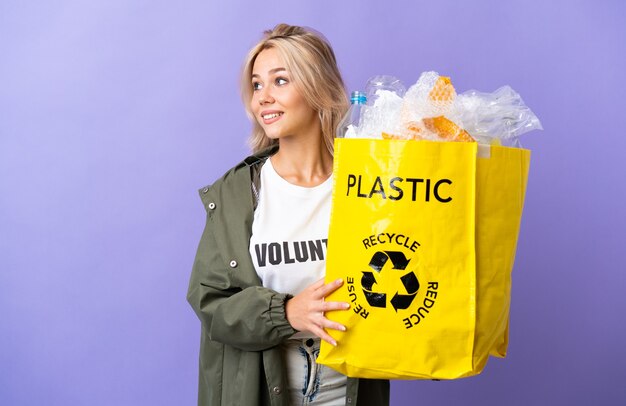 Jovem russa segurando uma sacola cheia de papel para reciclar isolada em roxo olhando para o lado