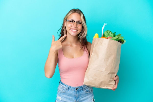 Jovem russa segurando uma sacola de compras de supermercado isolada em um fundo azul e fazendo um gesto de polegar para cima