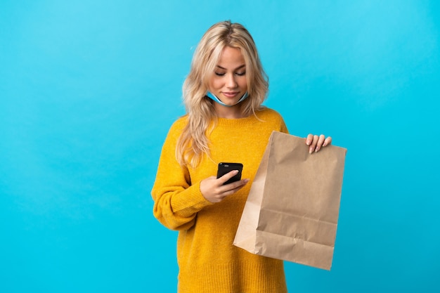 Jovem russa segurando uma sacola de compras de supermercado isolada em um fundo azul enviando uma mensagem com o celular