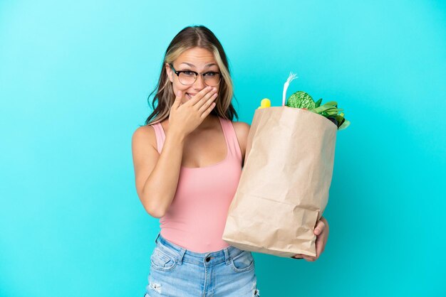 Jovem russa segurando uma sacola de compras de supermercado isolada em um fundo azul, feliz e sorridente, cobrindo a boca com a mão