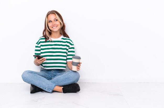 Jovem russa sentada no chão isolada em fundo branco segurando café para levar e um celular enquanto pensa em algo