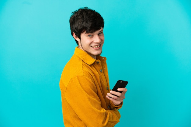 Jovem russo isolado em um fundo azul segurando um telefone celular e com os braços cruzados