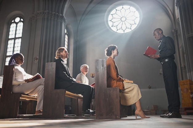 Foto jovem sacerdote africano lendo a bíblia para pessoas fiéis enquanto elas estão sentadas no banco da igreja