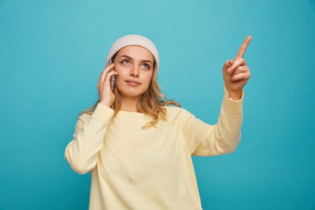 Jovem satisfeita com um chapéu de inverno falando no telefone, olhando e apontando para cima