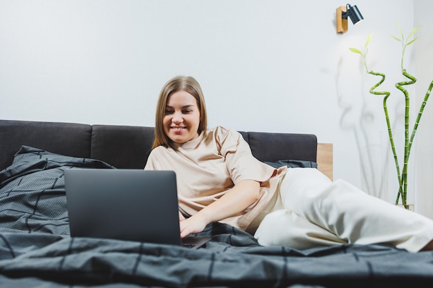 Jovem satisfeita terminando o trabalho no laptop Menina europeia sentada com o computador na cama estudando em casa ensino à distância e conceito de trabalho freelance remoto