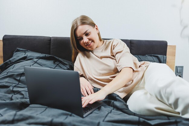 Jovem satisfeita terminando o trabalho no laptop Menina europeia sentada com o computador na cama estudando em casa ensino à distância e conceito de trabalho freelance remoto