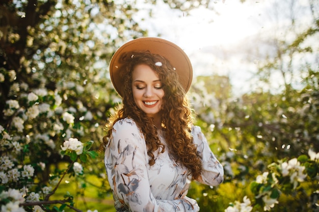 Jovem se divertindo entre as árvores florescendo em um dia de primavera. mulher de chapéu bege e vestido branco cheira a flor das flores nas árvores do jardim.