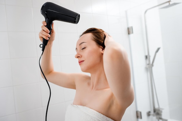 Jovem secando e estilizando o cabelo com secador de cabelo fazendo penteado no interior do banheiro moderno Rotina de beleza após o banho matinal