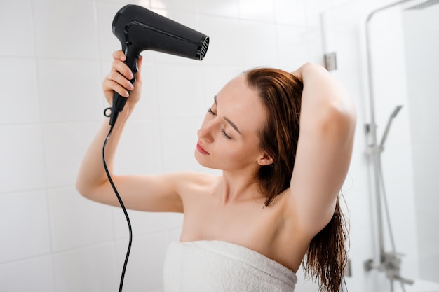 Jovem secando e estilizando o cabelo com secador de cabelo fazendo penteado no interior do banheiro moderno Rotina de beleza após o banho matinal