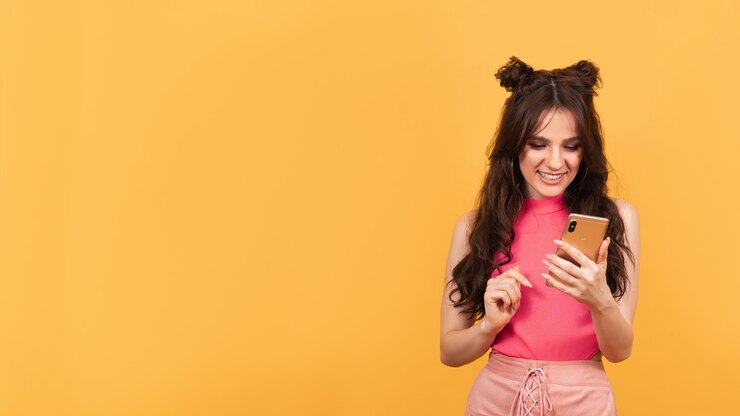Foto jovem segura o smartphone nas mãos e olha para a tela sorrindo em fundo laranja