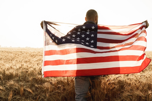 Jovem segurando a bandeira americana nas costas enquanto está em um campo de trigo