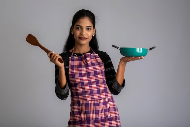 Jovem segurando e posando com uma espátula de utensílios de cozinha e uma panela em uma parede cinza