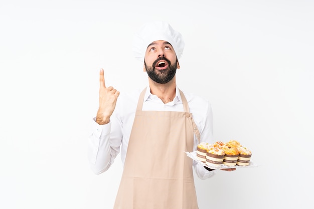 Jovem, segurando o bolo muffin sobre branco isolado surpreso e apontando para cima