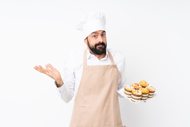 Jovem, segurando o bolo muffin sobre parede branca isolada, fazendo o gesto de dúvidas