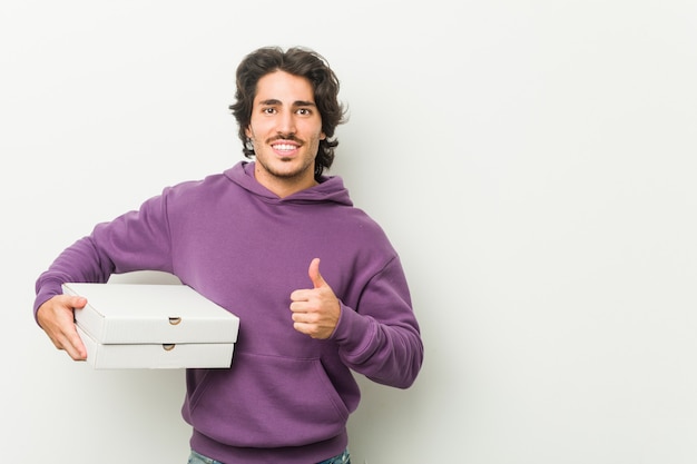 Jovem, segurando o pacote de pizzas, sorrindo e levantando o polegar