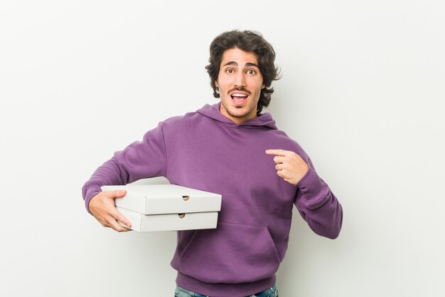 Jovem, segurando o pacote de pizzas surpreendeu apontando para si mesmo, sorrindo amplamente.