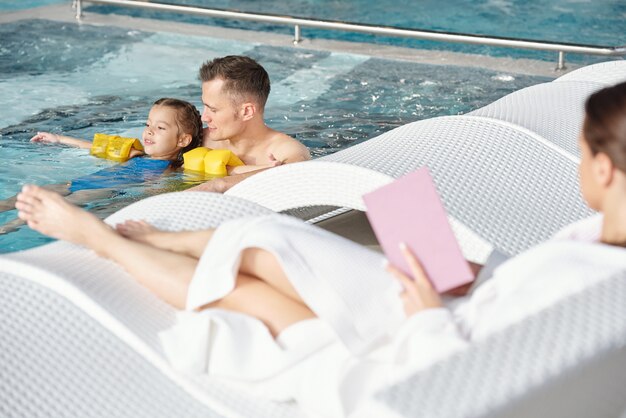 Jovem segurando sua filha com mangas de segurança durante a natação na piscina, enquanto passa um tempo no centro de spa no lazer