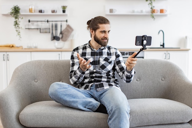 Jovem segurando telefone e selfie stick enquanto fala via webcam na cozinha