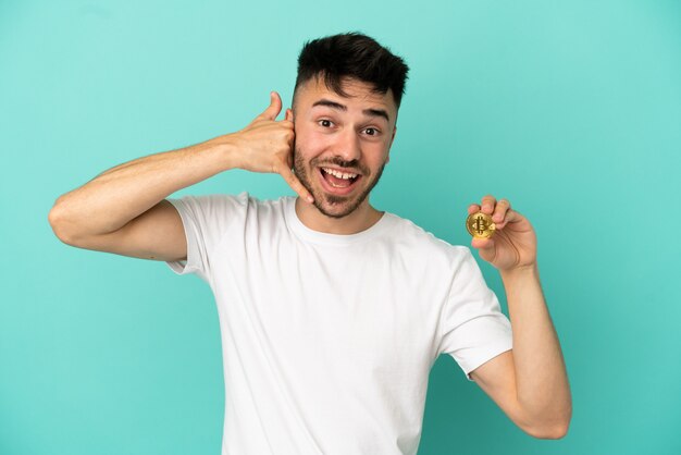 Jovem segurando um Bitcoin isolado em um fundo azul, fazendo gesto de telefone. Ligue-me de volta sinal