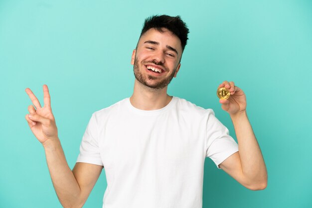 Jovem segurando um Bitcoin isolado em um fundo azul, sorrindo e mostrando sinal de vitória