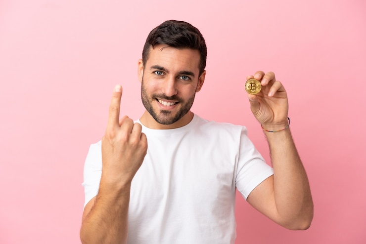 Foto jovem segurando um bitcoin isolado em um fundo rosa fazendo um gesto próximo