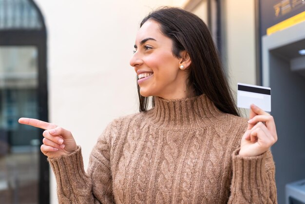 Jovem segurando um cartão de crédito ao ar livre apontando para o lado para apresentar um produto