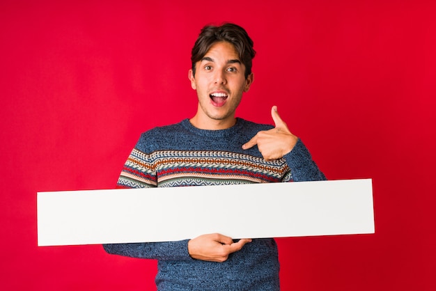 Jovem, segurando um cartaz surpreso apontando para si mesmo, sorrindo amplamente.