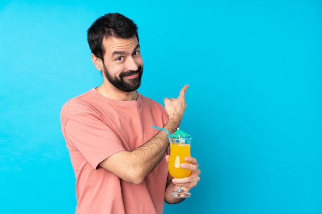 Jovem, segurando um cocktail sobre parede azul isolada, apontando para trás