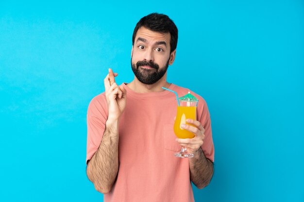 Jovem segurando um cocktail sobre parede azul isolada com dedos cruzando e desejando o melhor