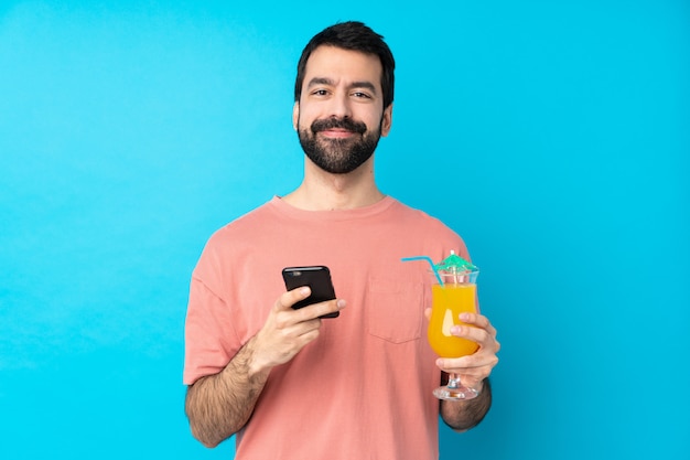 Jovem, segurando um cocktail sobre parede azul isolada, enviando uma mensagem com o celular