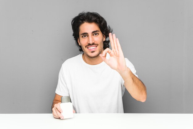 Jovem, segurando um creme pós-barba alegre e confiante mostrando okey gesto.