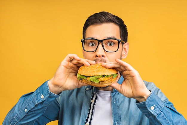 Jovem segurando um pedaço de sanduíche Estudante come fast-food Hambúrguer não é comida útil Cara com muita fome Conceito de dieta Isolado sobre fundo amarelo