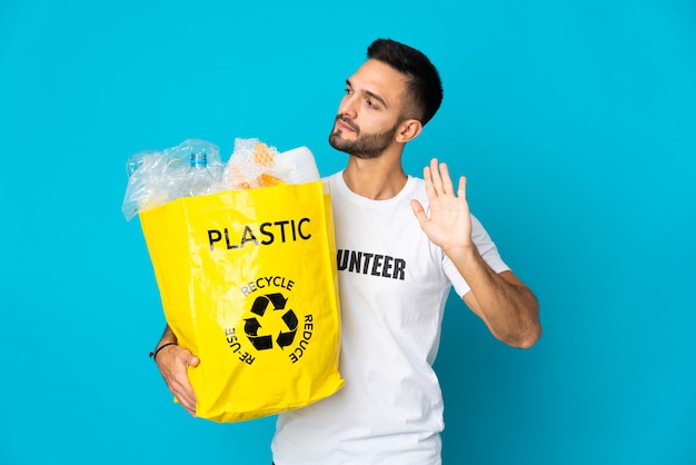 Jovem segurando um saco cheio de garrafas de plástico para reciclar isolado na parede azul fazendo gesto de pare e desapontado