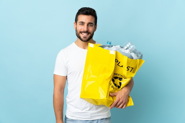 Jovem, segurando um saco cheio de garrafas de plástico para reciclar isolado na parede azul, sorrindo muito