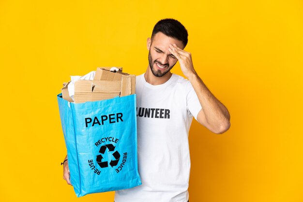 Jovem segurando um saco de reciclagem cheio de papel para reciclar isolado na parede branca com dor de cabeça