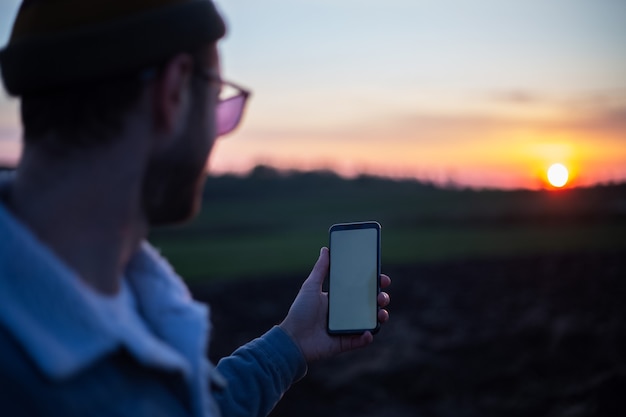 Jovem segurando um smartphone com uma maquete branca vazia no fundo do pôr do sol