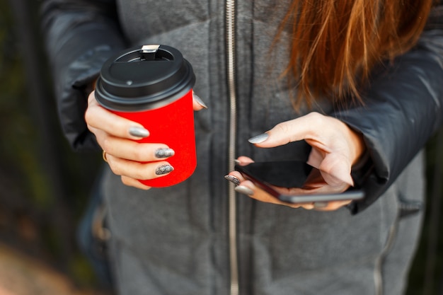 Jovem segurando um smartphone preto e bebendo um café delicioso ao ar livre Close de mãos femininas