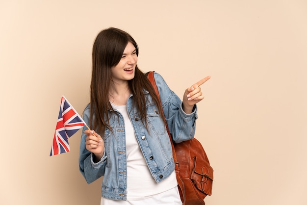 Jovem segurando uma bandeira do Reino Unido isolada em um bege, apontando o dedo para o lado e apresentando um produto