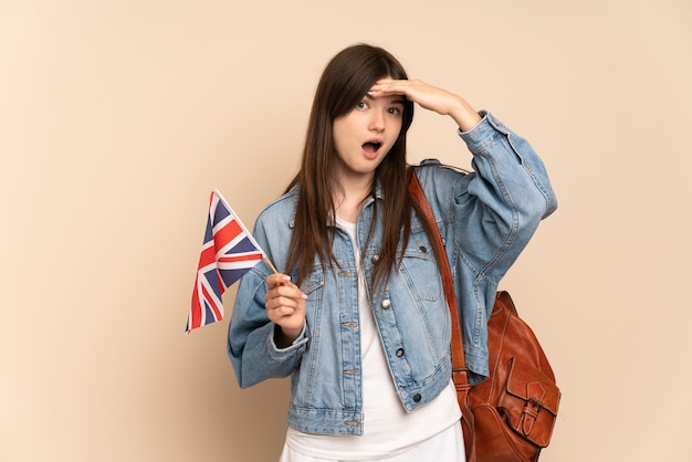 Jovem segurando uma bandeira do Reino Unido, isolada em um tom bege, fazendo um gesto surpresa enquanto olha para o lado