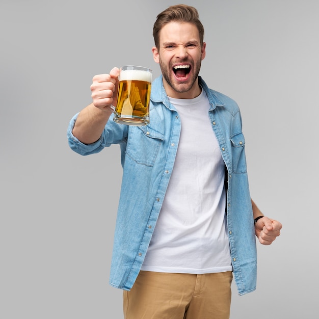 Jovem segurando uma camisa jeans segurando um copo de cerveja em pé sobre a parede cinza.