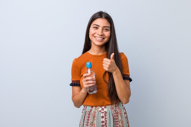 Jovem, segurando uma garrafa de água, sorrindo e levantando o polegar