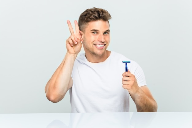 Jovem, segurando uma lâmina de barbear, mostrando sinal de vitória e sorrindo amplamente.