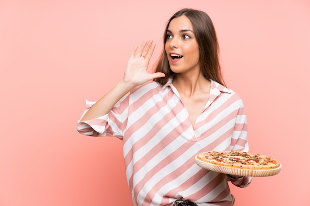 Foto jovem, segurando uma pizza sobre parede rosa isolada, gritando com a boca aberta