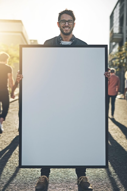 Foto jovem segurando uma placa de sinal vazia ou cartaz enquanto está do lado de fora na rua da cidade retrato de um cara feliz, sorridente e alegre, fazendo um anúncio mostrando um cartaz para promoção