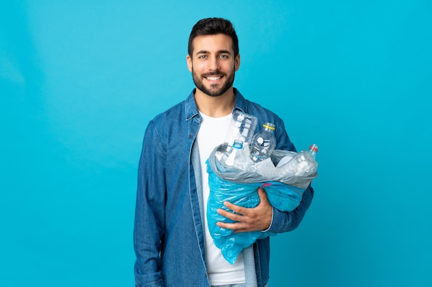 Jovem segurando uma sacola cheia de garrafas plásticas para reciclar isoladas na parede azul sorrindo muito